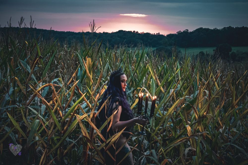 Woman with dark hair holding glowing candles in a cornfield at dusk with a dramatic purple sunset sky.