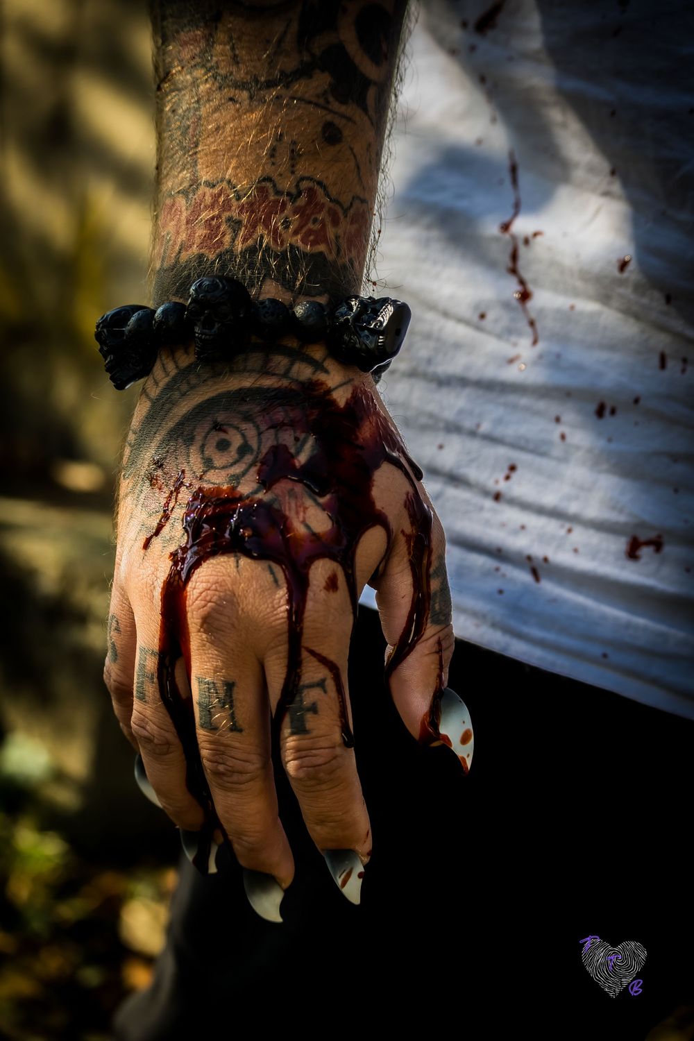 Tattooed hand with Halloween costume makeup bloody wound effect, black bead bracelet, white shirt with fake blood spatters.