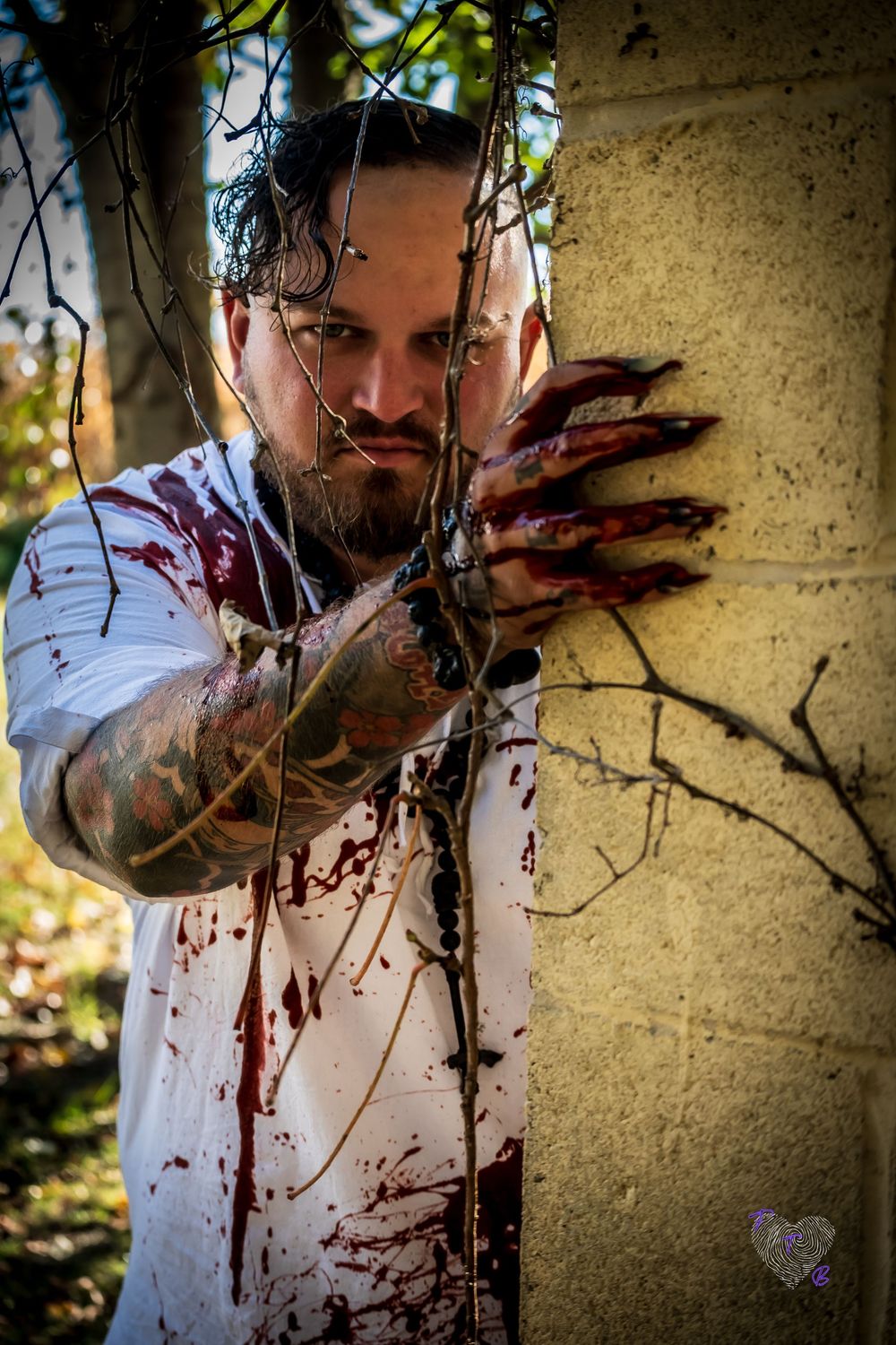 Tattooed man in bloody white shirt clutches a stone pillar with clawed hands amid dark branches in a horror-themed photo.