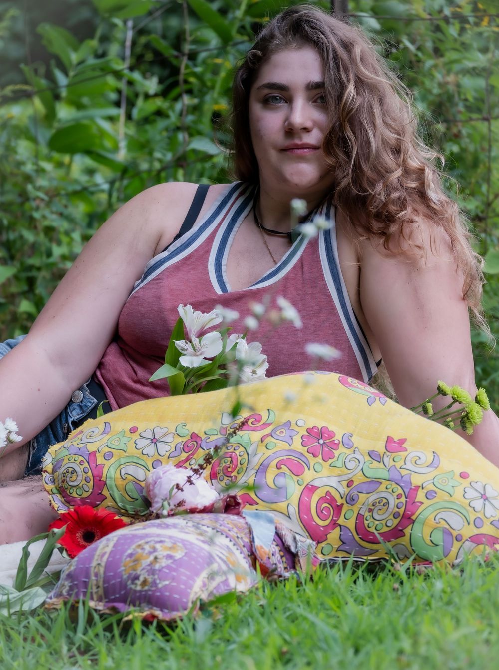 A figure in a red tank top with curly hair relaxes in the grass with a colorful paisley pillow.