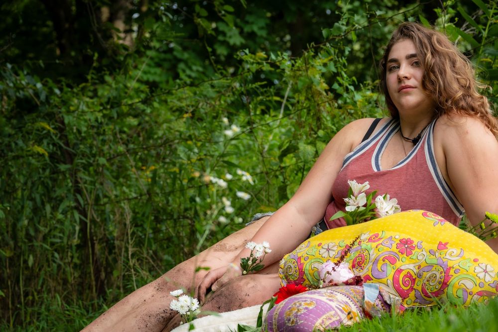 A person in colorful summer attire reclines outdoors in lush green grass holding white wildflowers.
