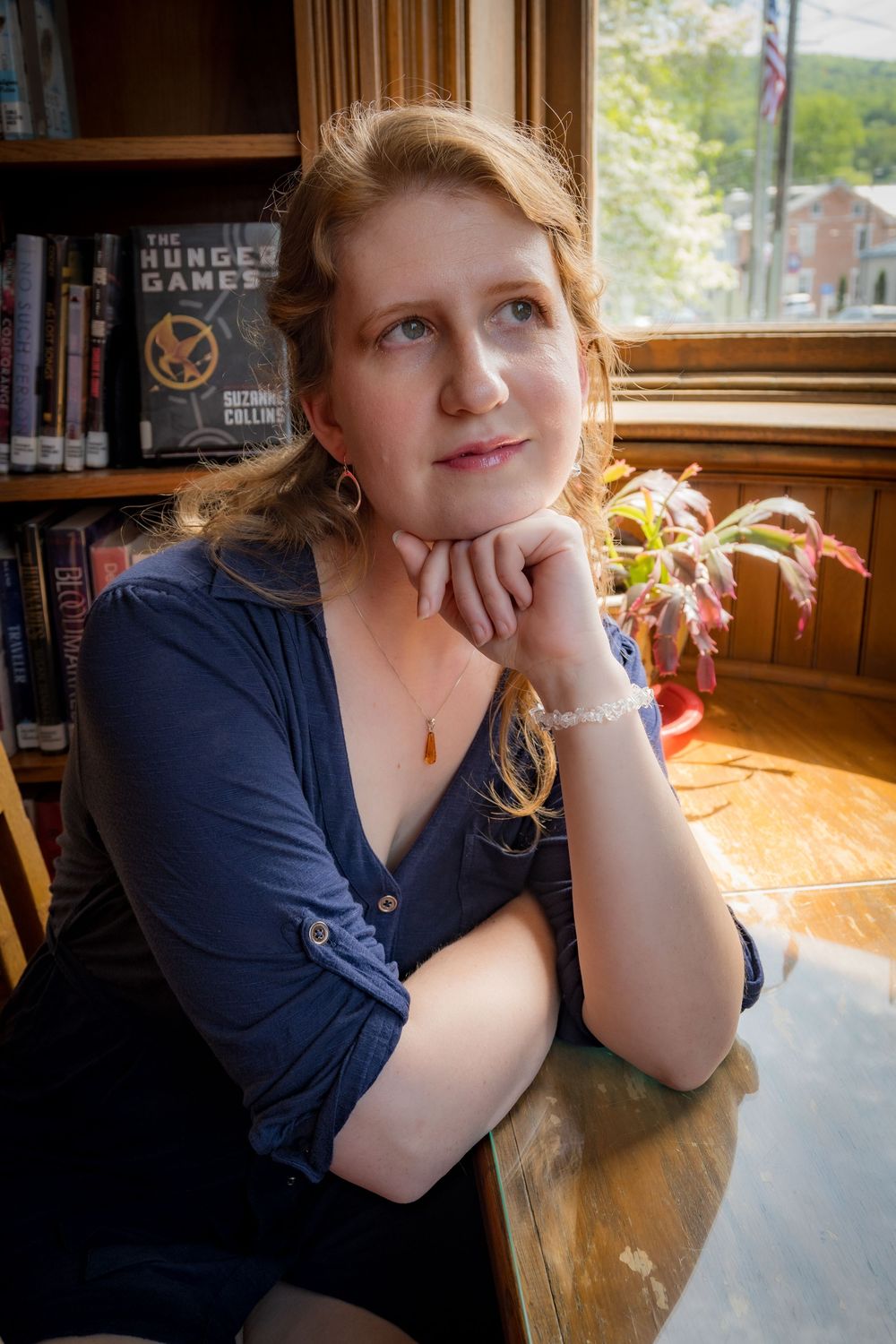 Interior photo with warm lighting shows someone seated by a window with bookshelves in the background.