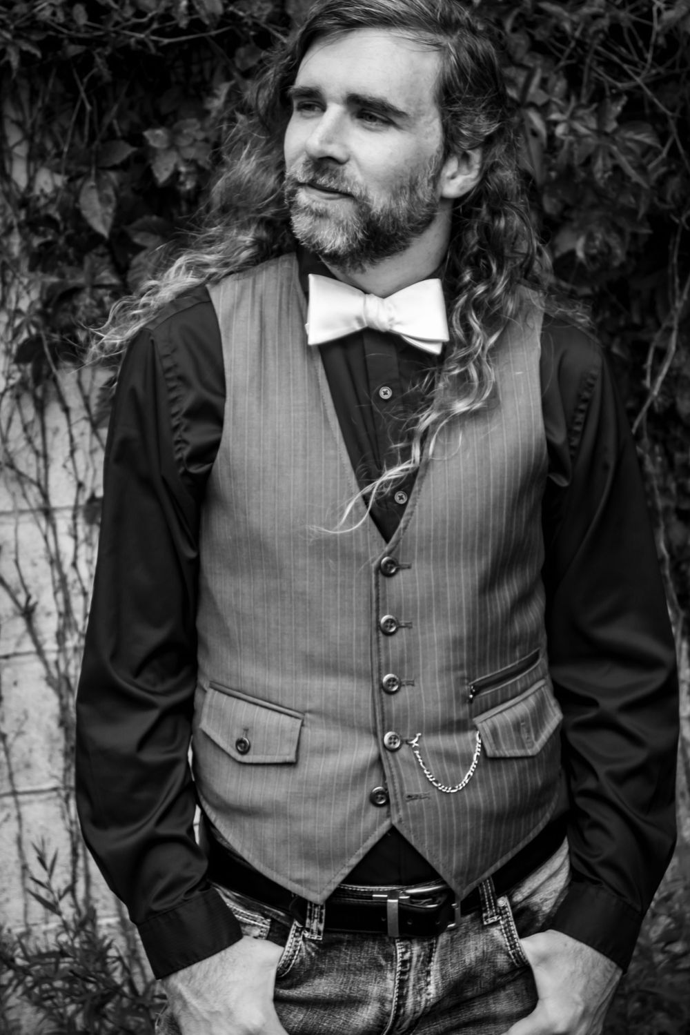 Black and white portrait of a long-haired bearded man in a vest, bow tie, and jeans posing outdoors.