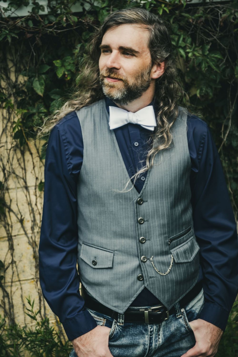 Bearded man with long curly hair wearing a gray vest, bow tie, and jeans, posing outdoors near green ivy.