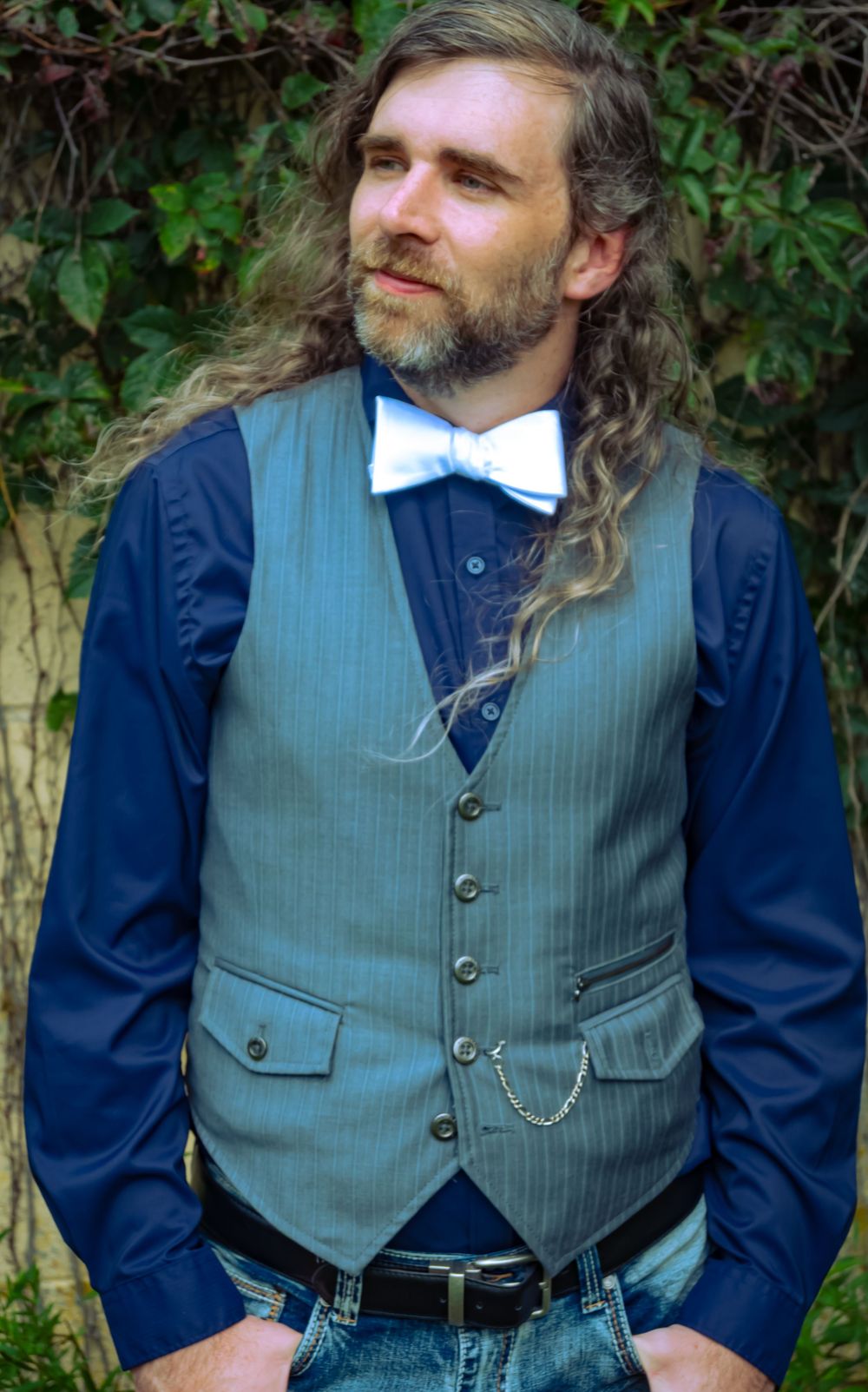 Man with long hair and beard wearing a gray vest, white bow tie, and navy shirt, posing outdoors.
