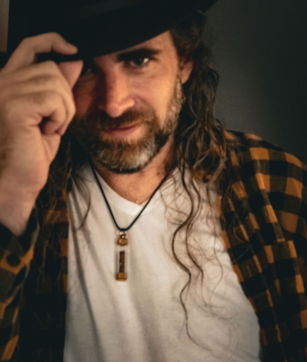 Man with long curly hair, beard, black cowboy hat, plaid shirt, and pendant necklace posing confidently.
