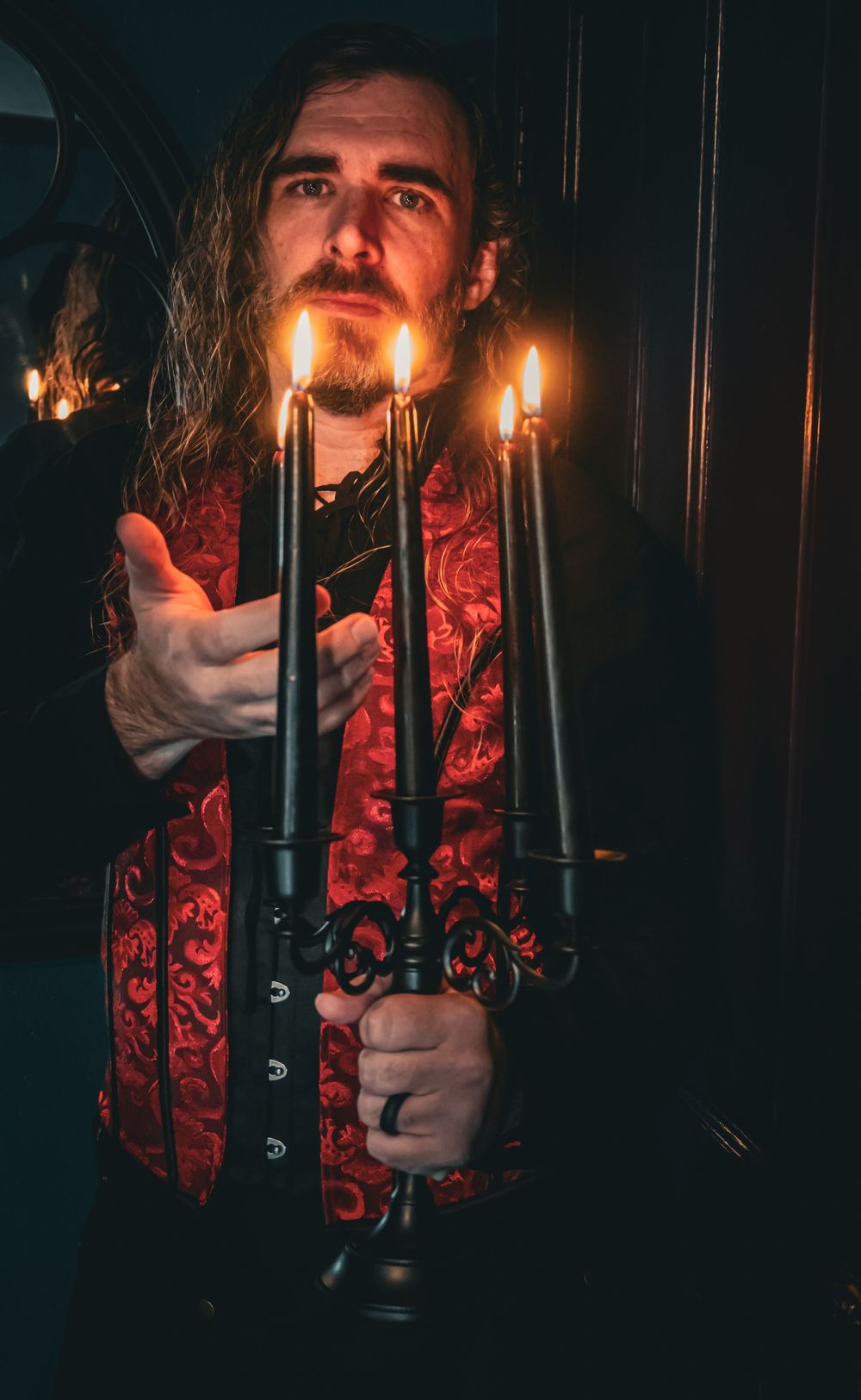 Man with long hair holding a black candelabra with lit candles, wearing a red brocade vest in a dark setting.