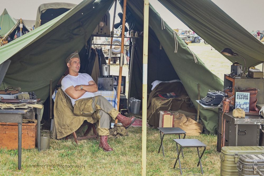 Military reenactor rests in a vintage canvas tent with period equipment and furniture at an outdoor encampment.