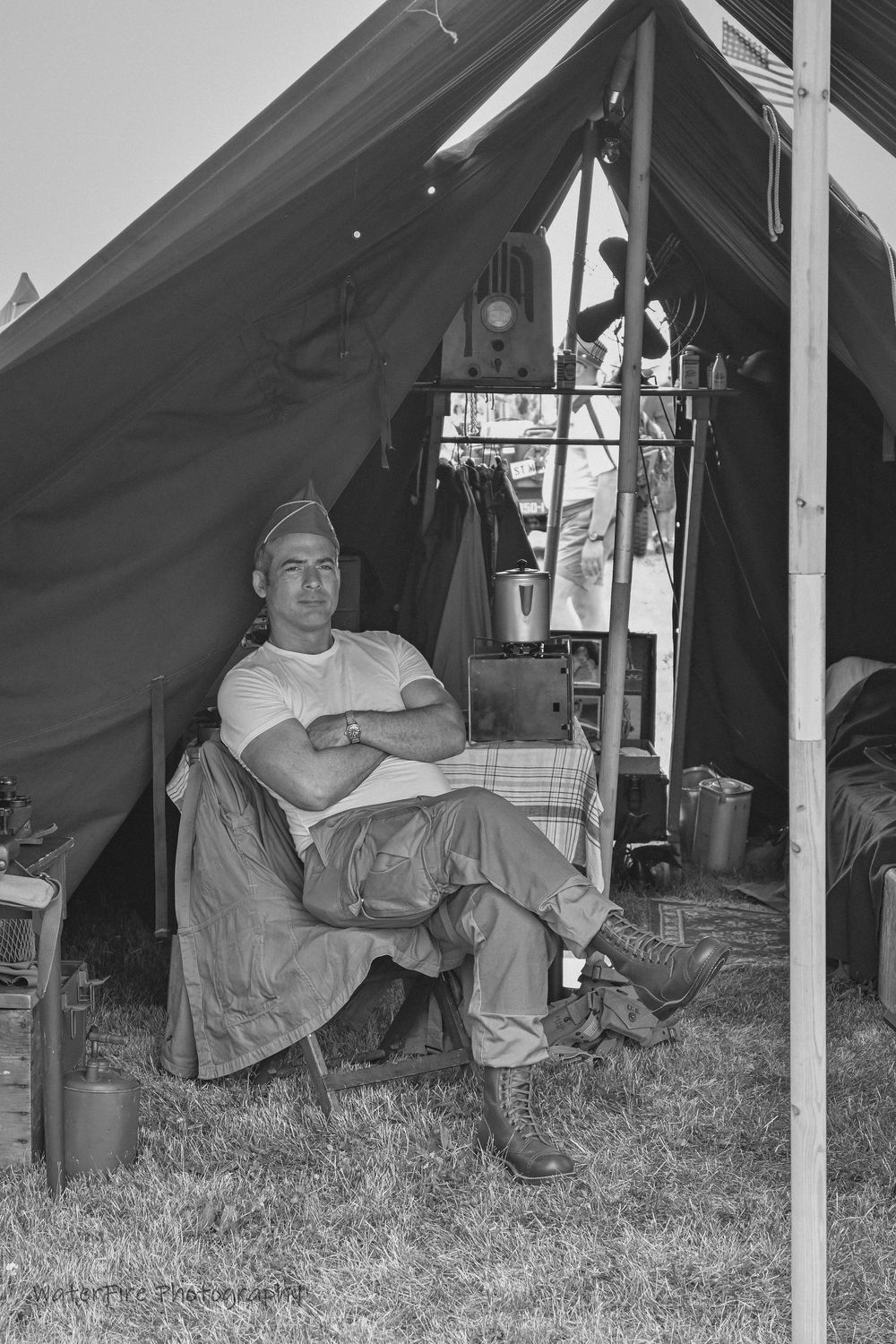 Soldier in uniform relaxes in a chair at a military camp tent site during World War II, captured in black and white.