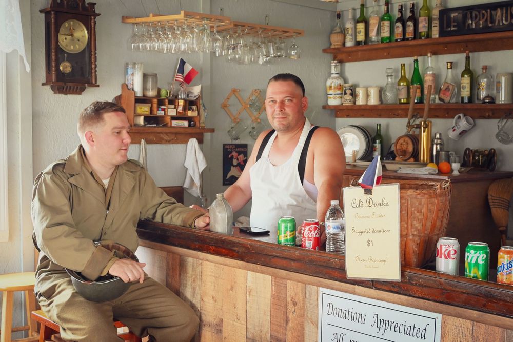Inside a rustic bar setting with liquor bottles and glassware on shelves, a customer in military attire chats with the bartender.