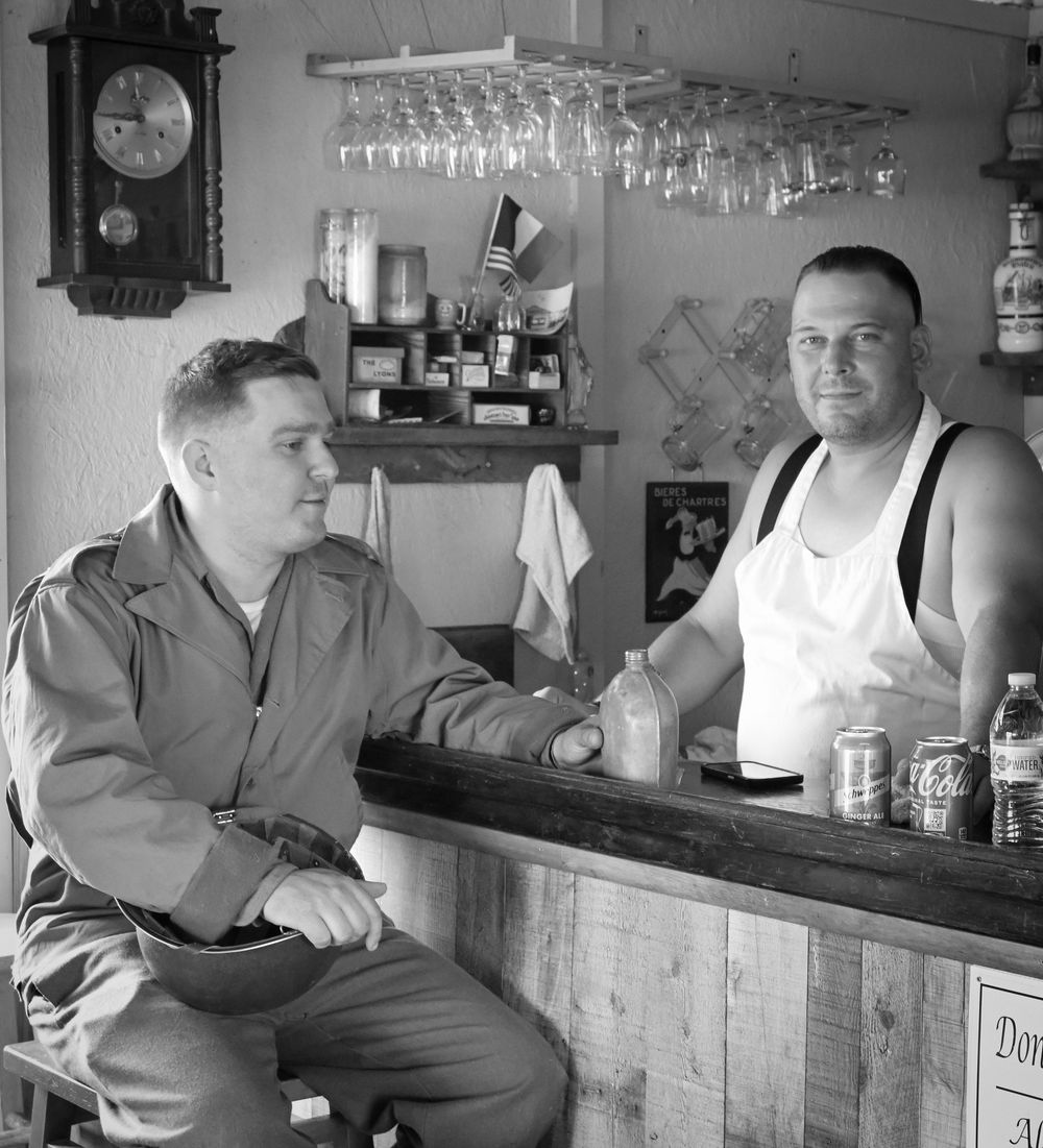 Black and white photo of a 1950s bar interior with two men, a clock on wall, and wine glasses hanging overhead.