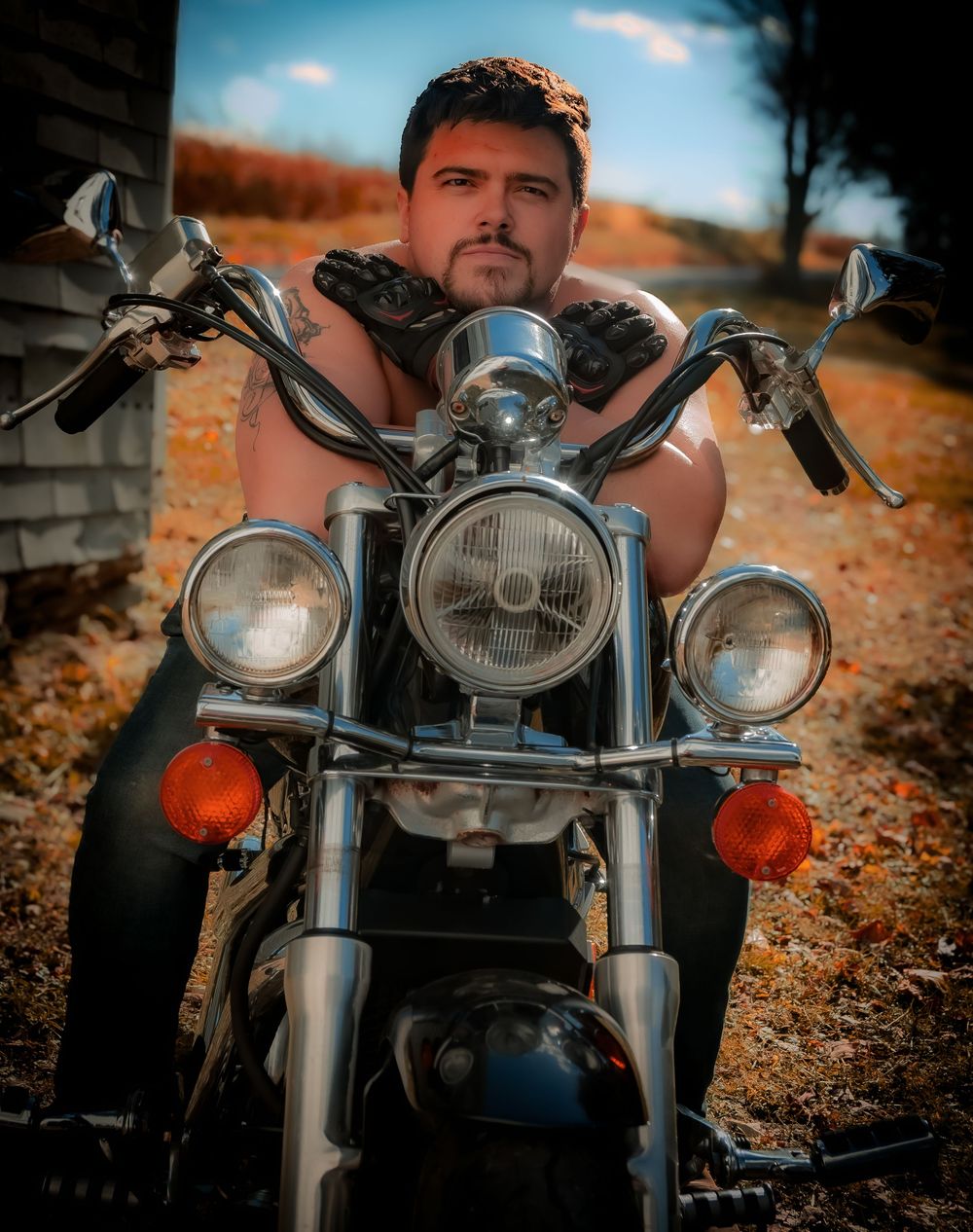Shirtless man with tattoos posing on a classic chrome motorcycle outdoors in autumn.