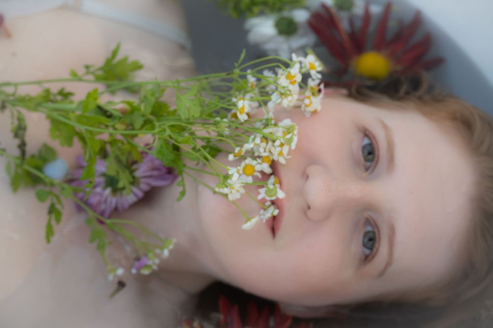 Young woman lying with wildflowers including chamomile and daisies arranged near her face in a serene floral portrait.