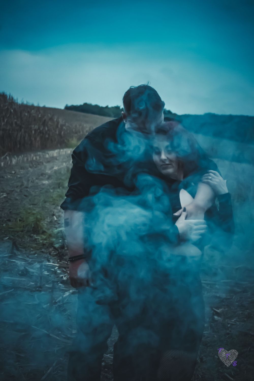 Couple embracing in a field surrounded by blue smoke under a moody sky.