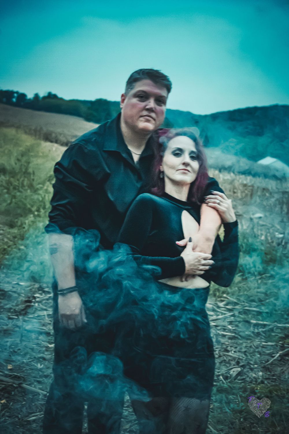 A couple embraces in a misty field, wearing black outfits, with colorful smoke swirling around them at dusk.