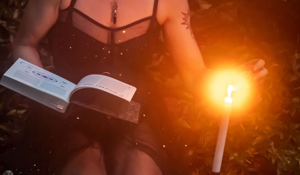 A person in dark clothing reads a book by glowing candlelight in a moody atmospheric setting.