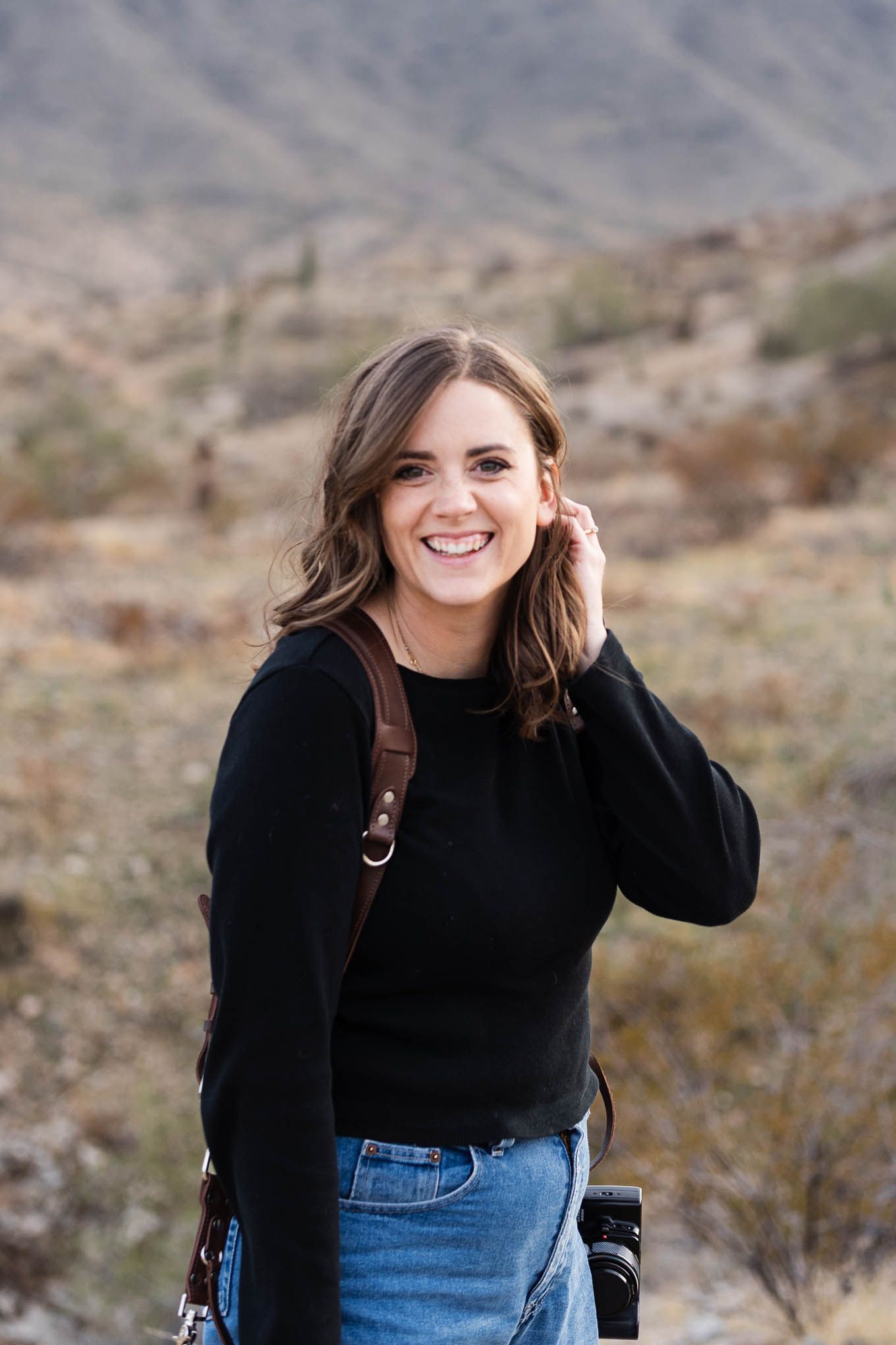 A person in a black sweater smiles while hiking in a desert mountain landscape.