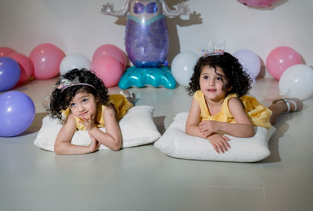 Two children lying on white pillows with pink and purple balloons and a metallic balloon decoration in the background.