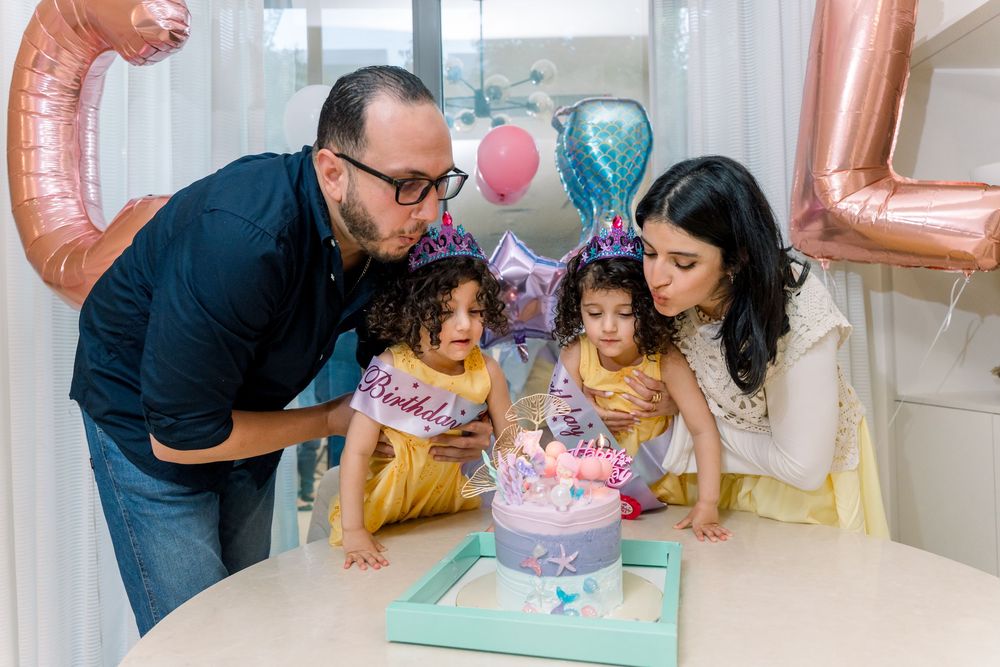 A family gathers around a birthday cake on a table with colorful balloons in the background at a celebration.