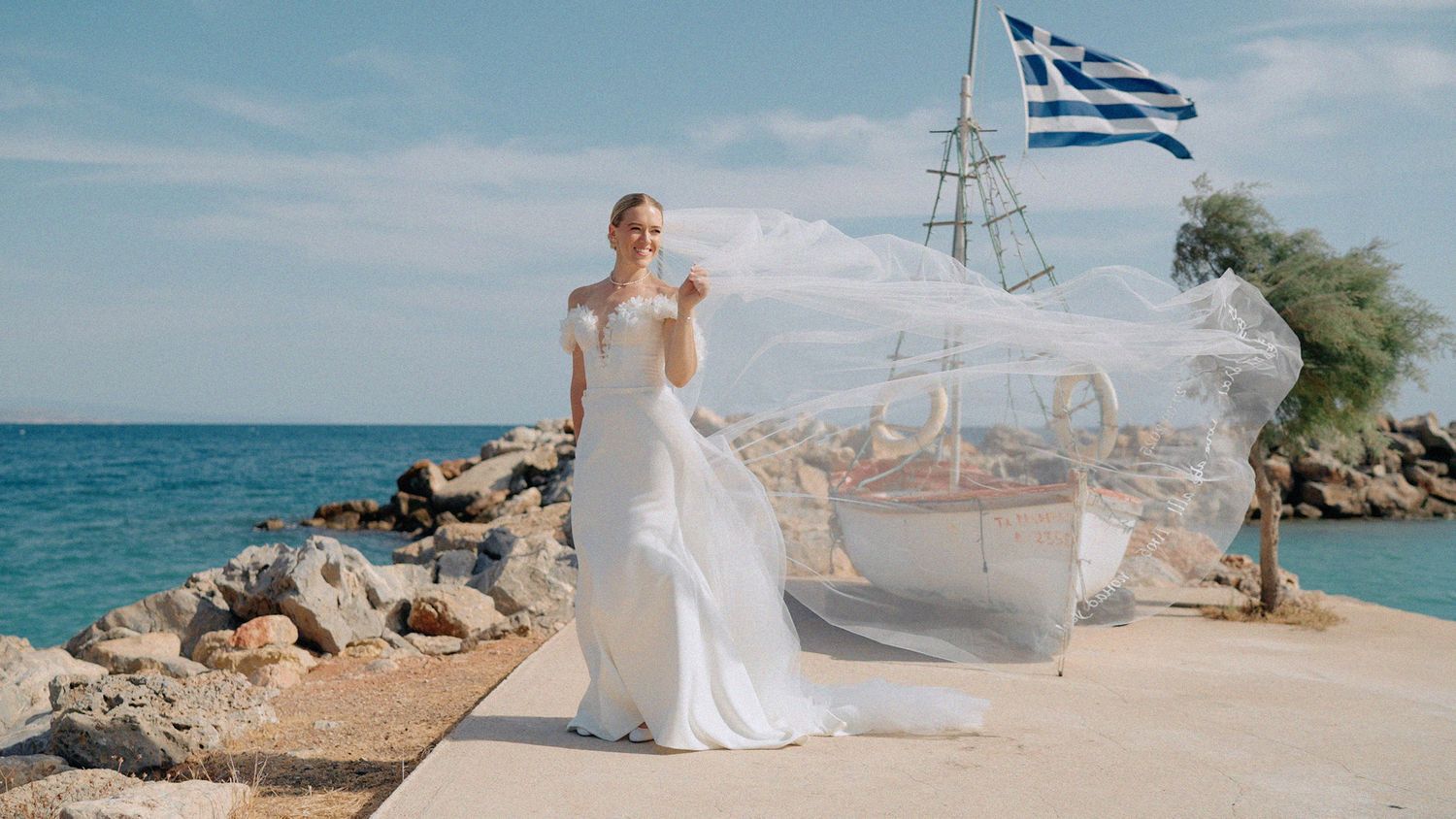 Wedding Videographer Chios
Bride in white wedding gown with flowing veil poses on rocky seaside pier with Greek flag waving in background.