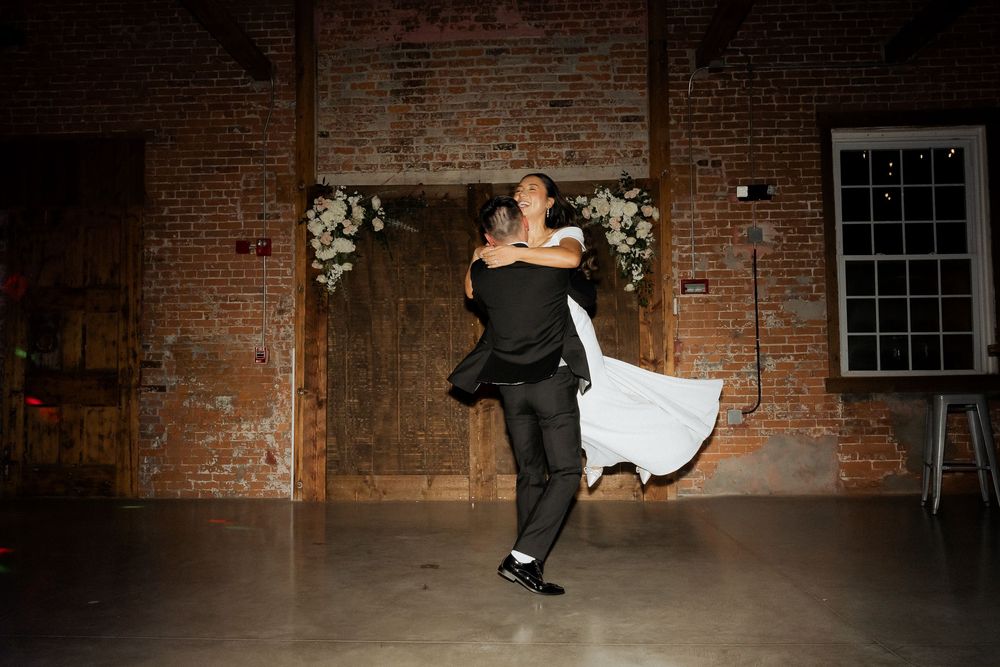 Bride and groom share a romantic first dance in a rustic brick venue adorned with white floral arrangements.