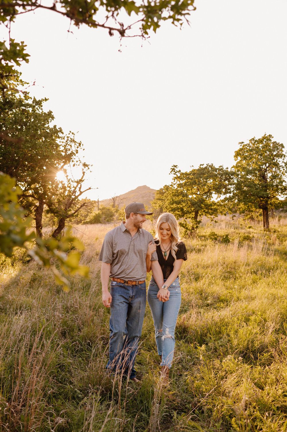 Couple walking through sunlit field with mountains in background during golden hour. Oklahoma Wedding Photographer