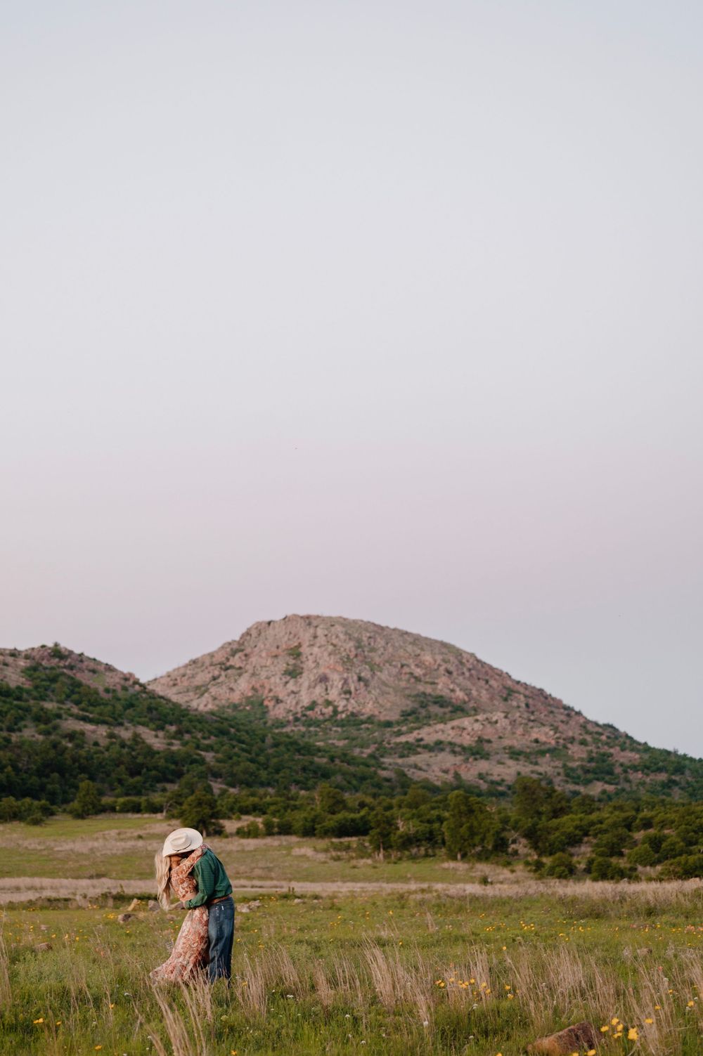 Scenic mountain landscape with couple embracing in distant wildflower field. Oklahoma Wedding Photographer