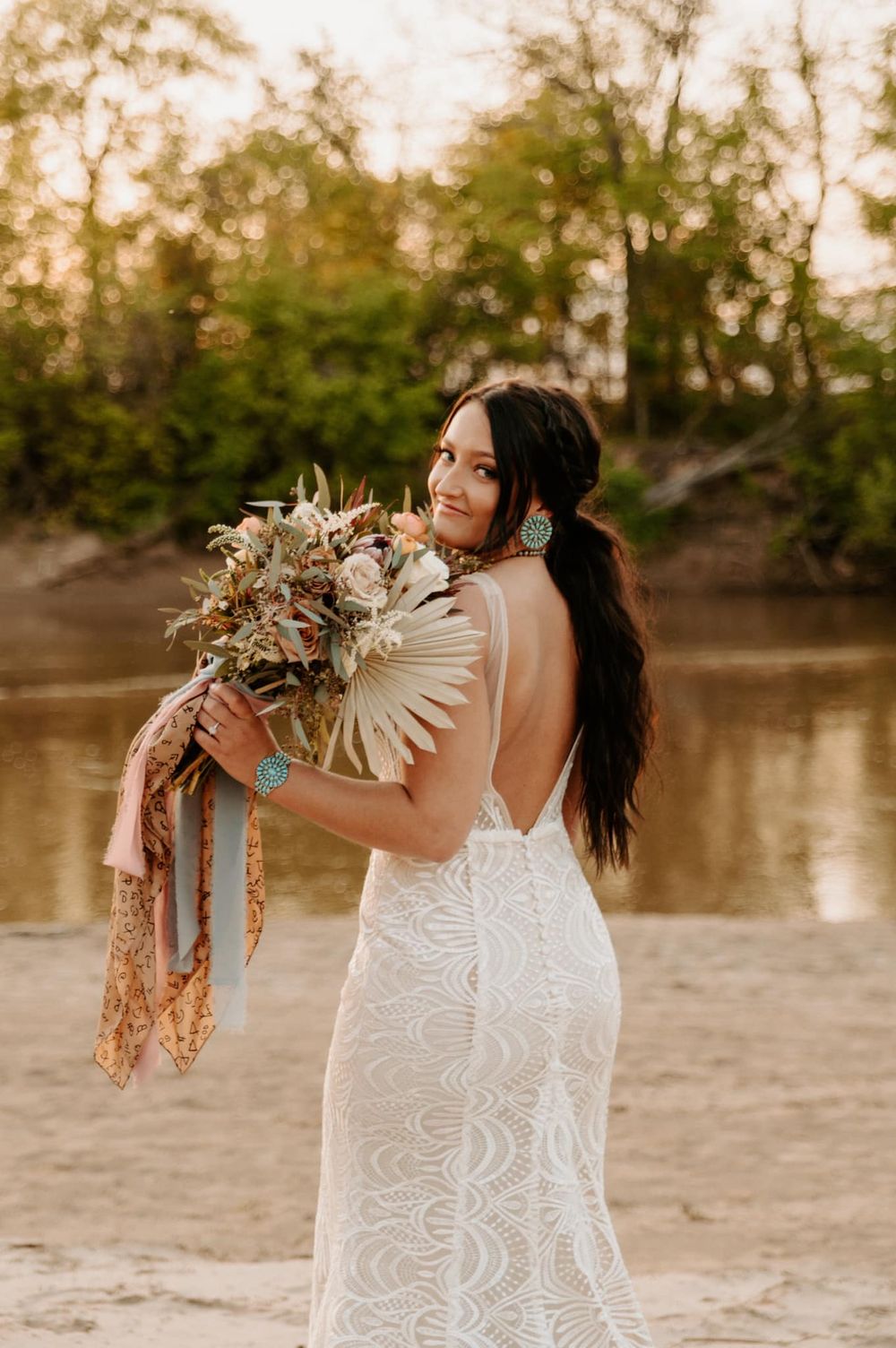 A bride in a lace dress holds a boho-style bouquet near a lake at sunset. Oklahoma Wedding Photographer