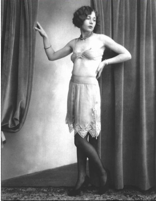Vintage 1920s boudoir photo featuring a woman in lingerie and lace skirt posing against curtains.