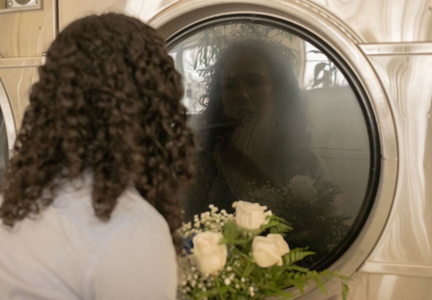 Person with curly hair holding white roses reflected in a washing machine door at a laundromat.