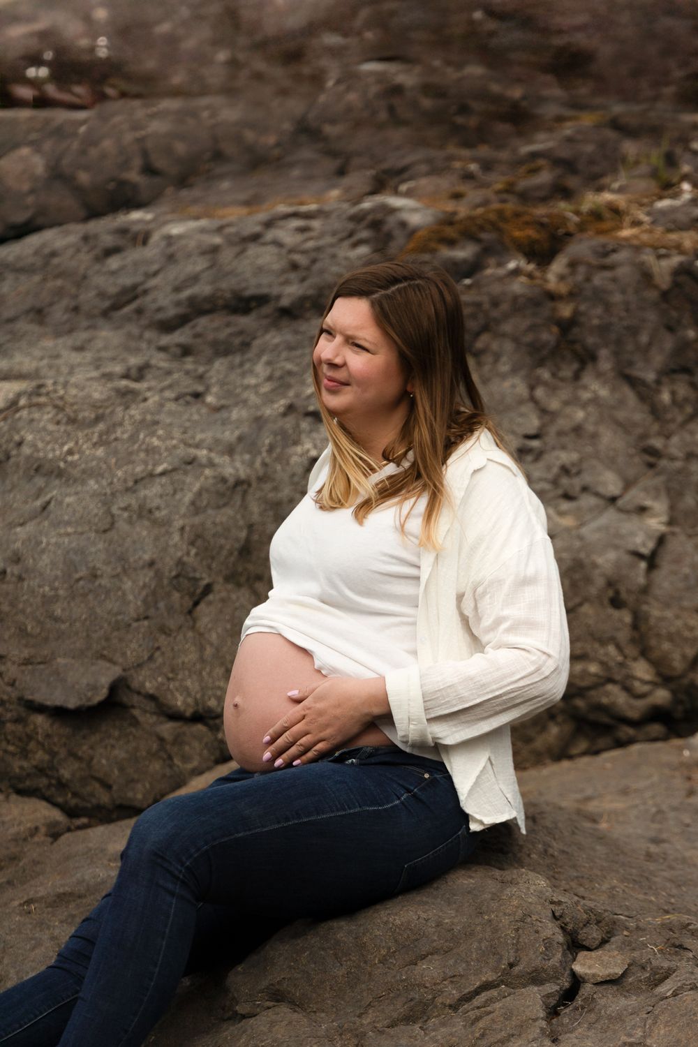Expectant mother in white cardigan sits on rocky outcrop during maternity photoshoot.