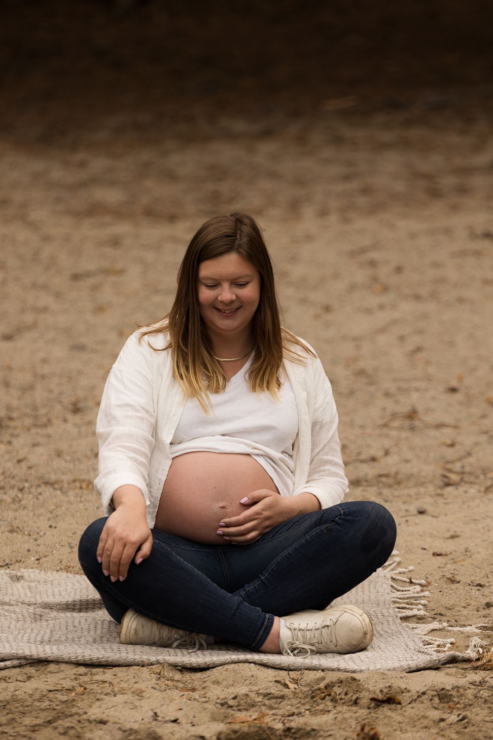 Maternity portrait of woman in white sitting cross-legged on blanket outdoors.