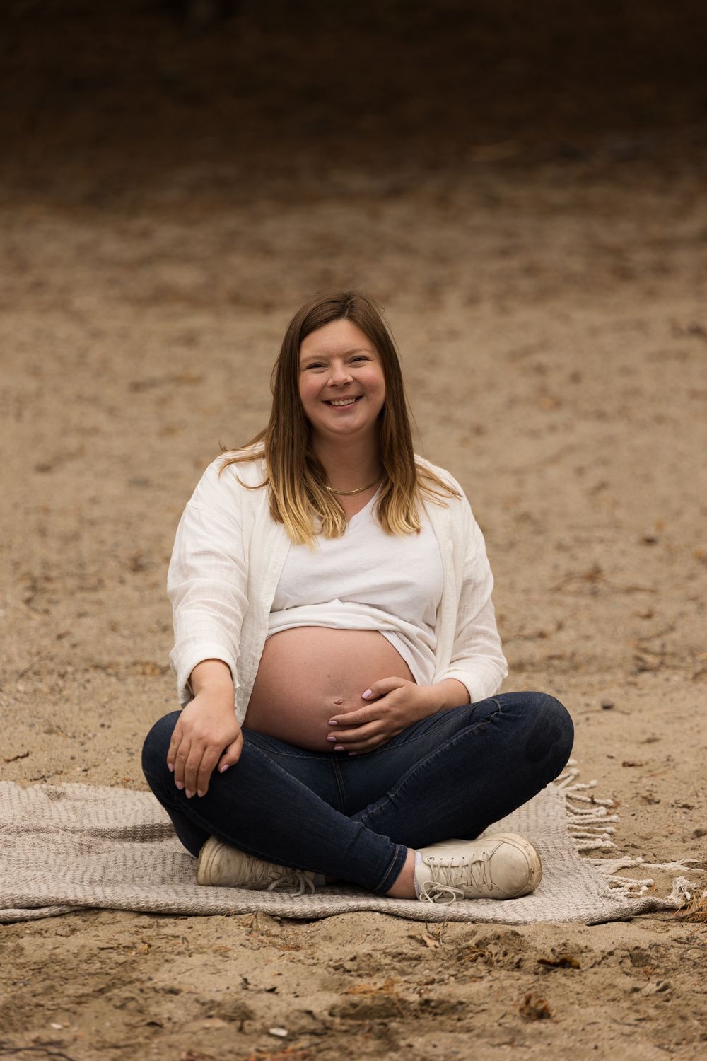 Maternity portrait of woman in white sweater sitting on blanket at beach.
