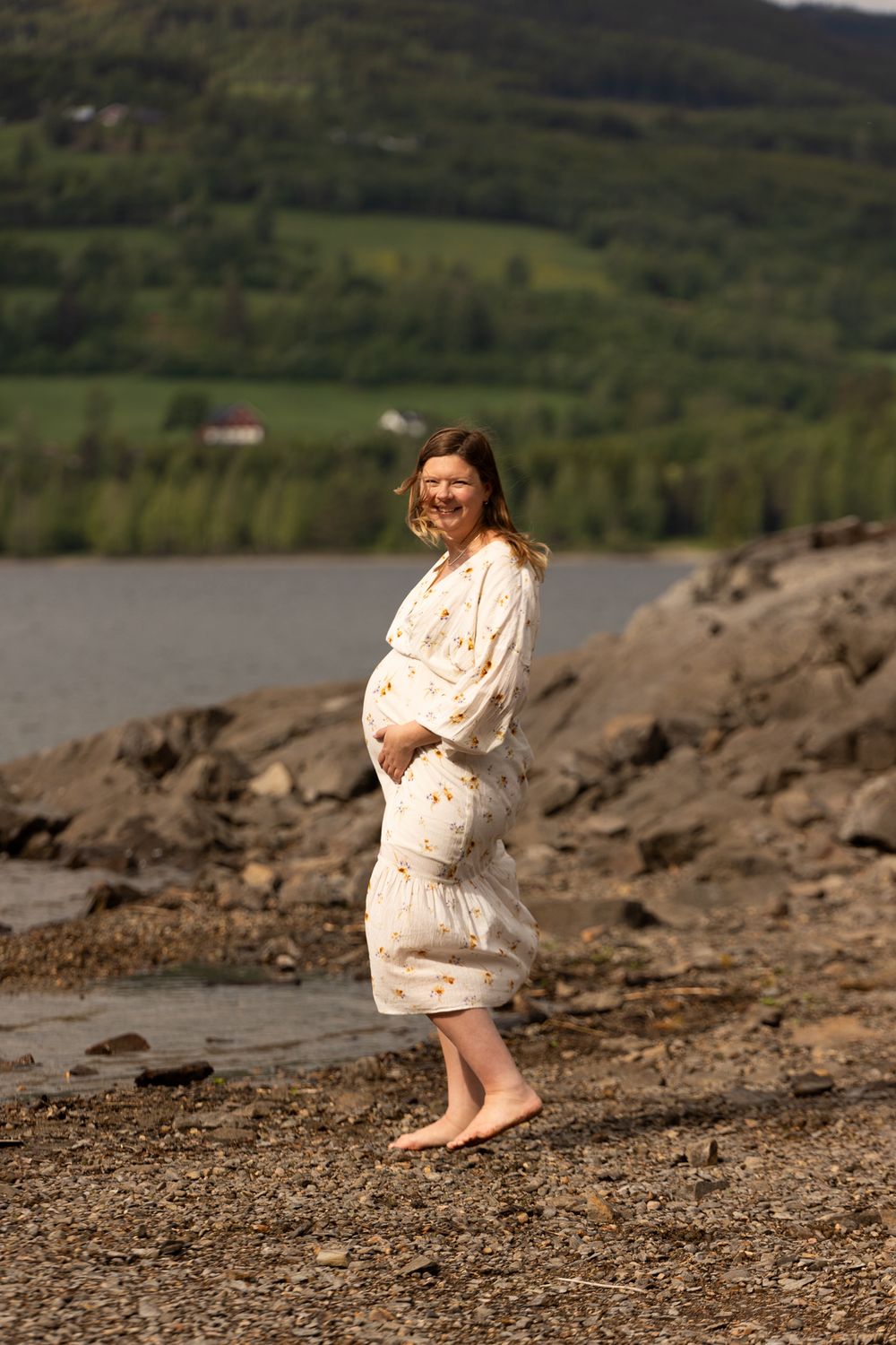 Maternity photo of woman in flowy dress standing barefoot by rocky shoreline.