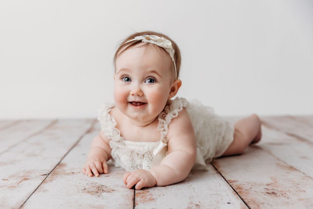 Baby in white lace outfit poses on floral backdrop during milestone photo session.
