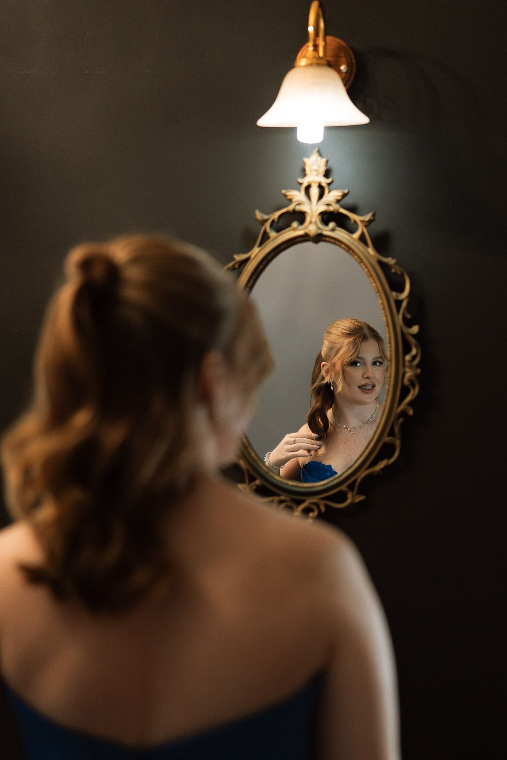 Woman in blue dress adjusts necklace while gazing at her reflection in an ornate gold mirror lit by a wall sconce.