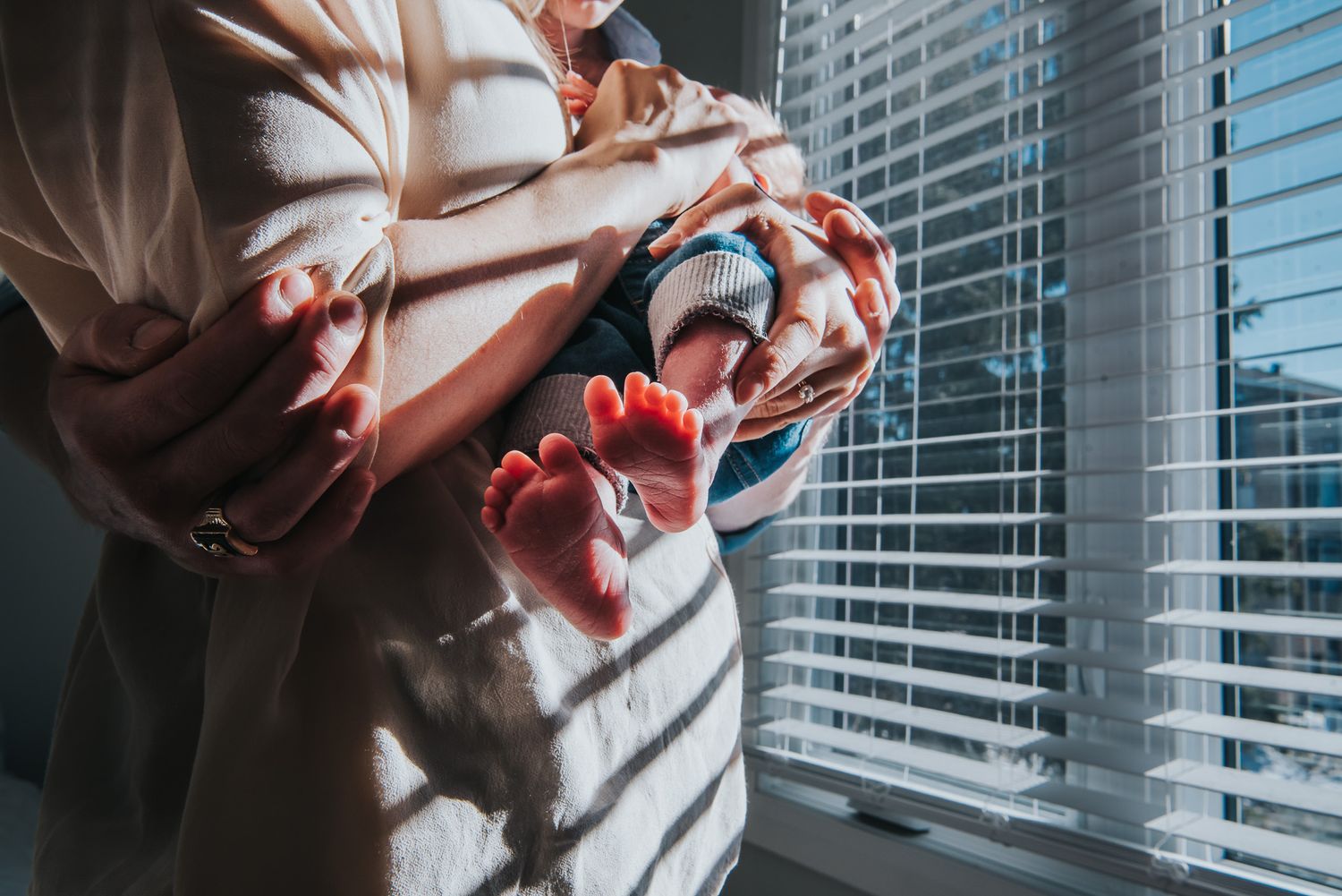 Colour image focused on a newborns feet as he's held by his parents in front of the window.