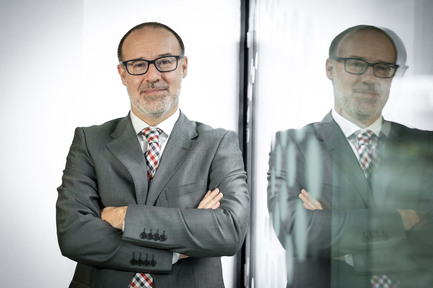 Profesional con traje gris, gafas y corbata a rayas rojas con los brazos cruzados junto a una superficie reflectante.