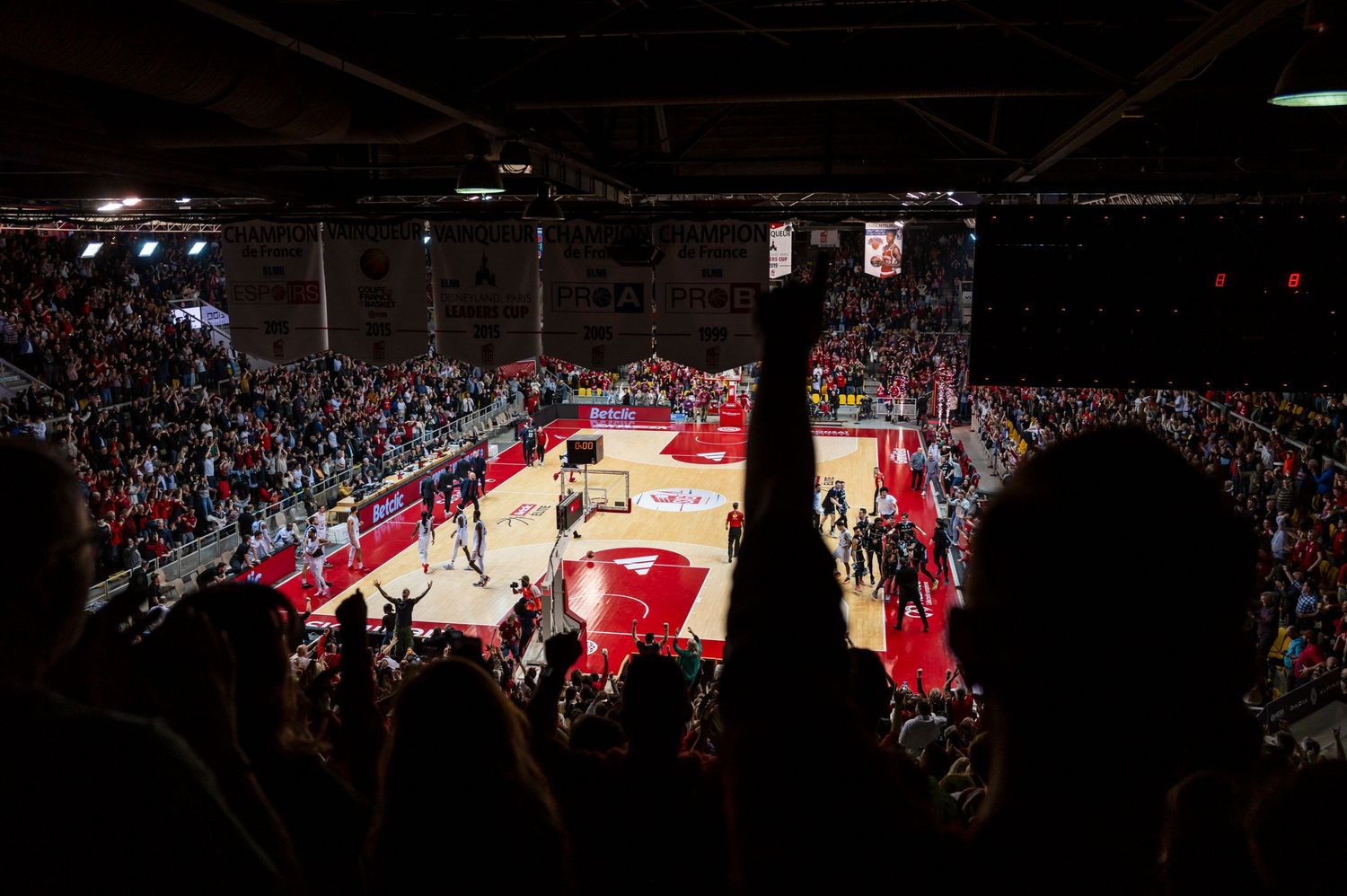 Basketball game in progress viewed from dark stadium seats with crowd watching.