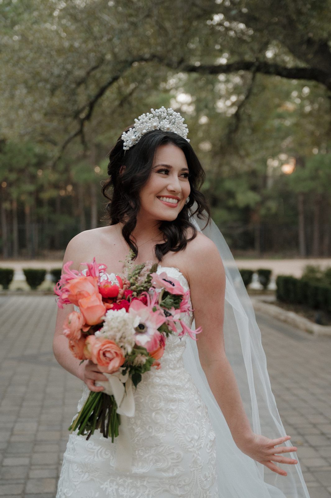 A bride in a white lace dress holds a vibrant coral and pink bouquet while posing on a brick patio surrounded by greenery.