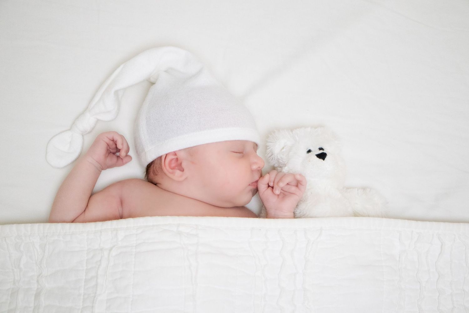 Newborn baby peacefully sleeping under white covers, cuddling a teddy bear. The scene has a serene atmosphere.