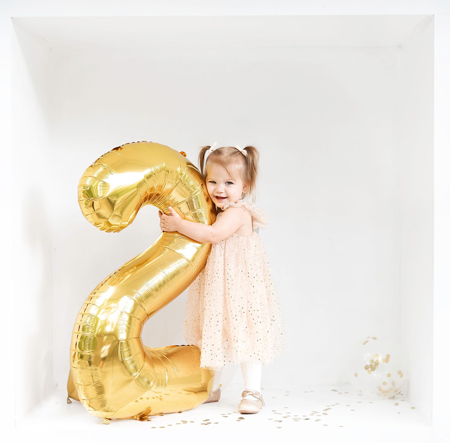 Small child in peach dress poses with large gold number 2 balloon against white background.