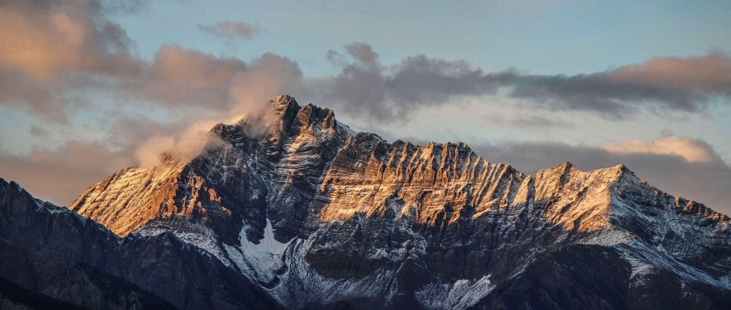Snow-capped mountain peak at sunset with dramatic clouds and warm golden light.