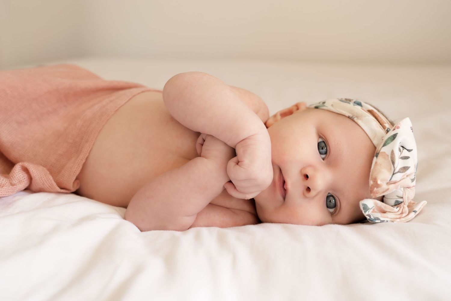 A peaceful portrait of an infant wearing a delicate white headband lying on white bedding.