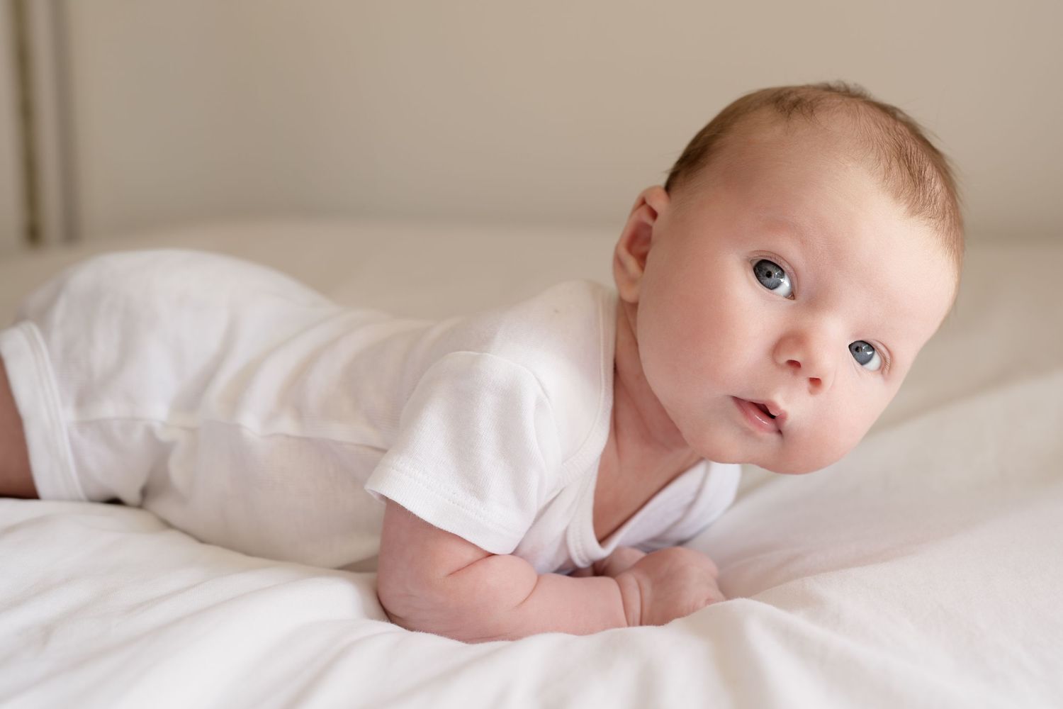 A bright portrait of an infant in white clothing propped on white pillows.