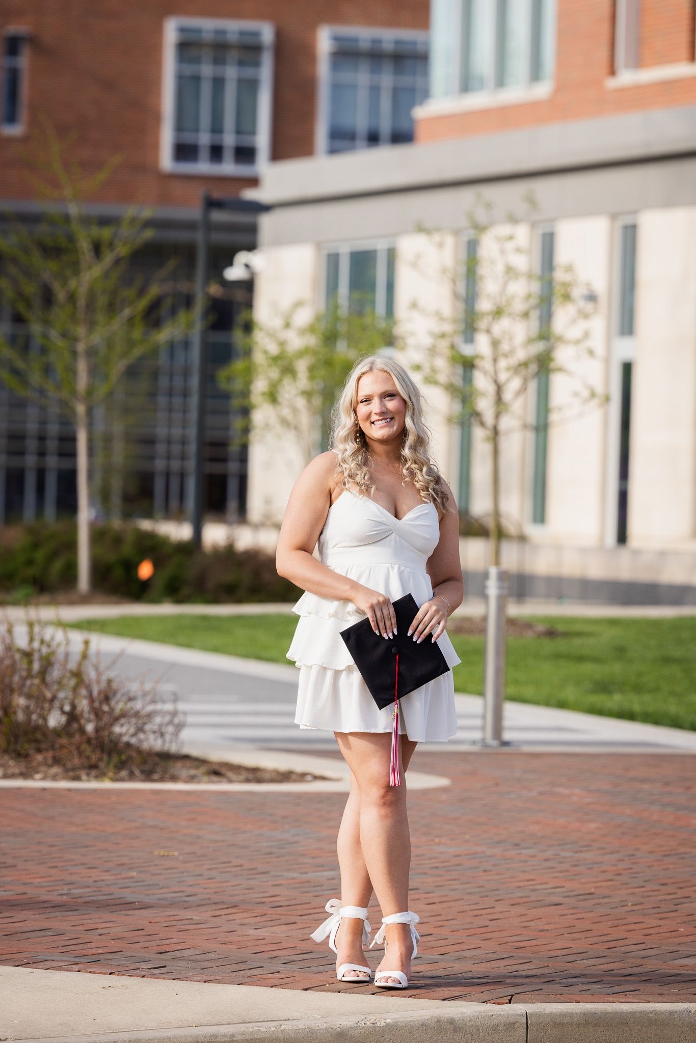 Student posing for graduation photos on brick pathway near university buildings.