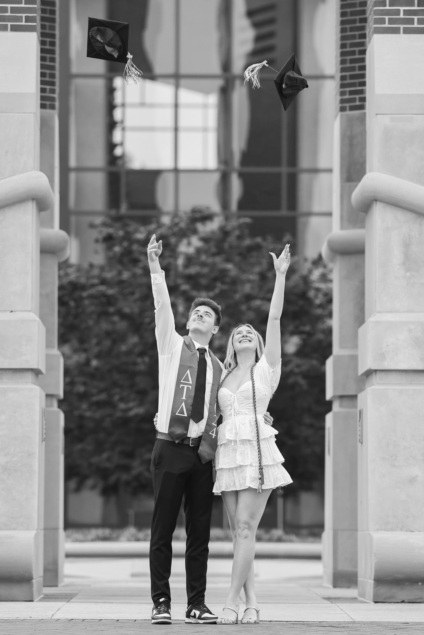 Graduates toss their caps in celebration between architectural pillars in black and white.