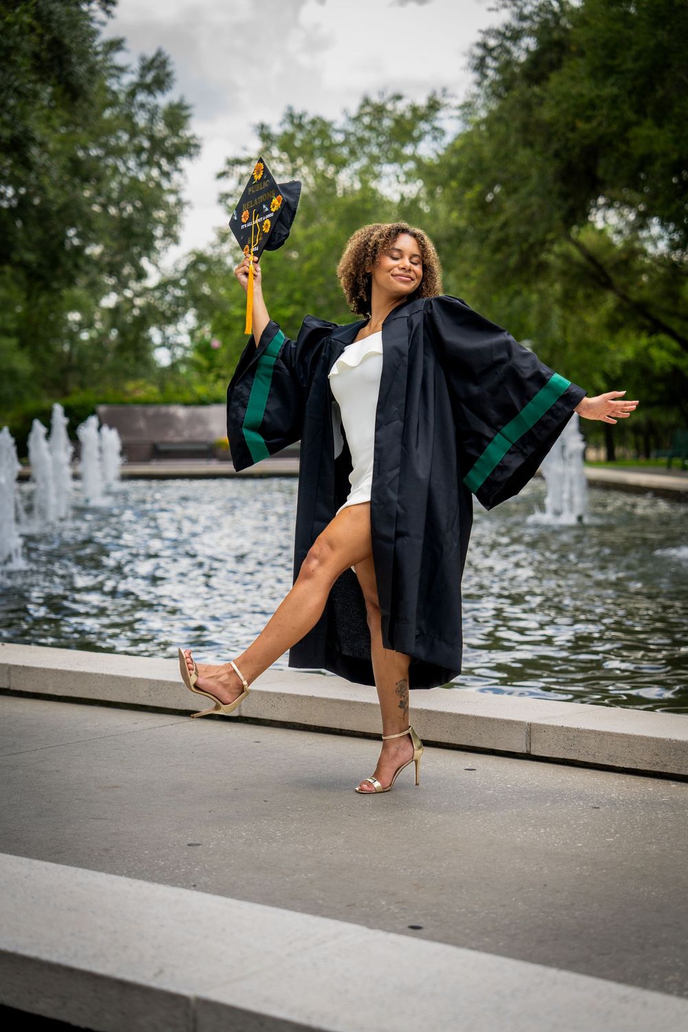 Joyful graduate in black cap and gown celebrating by fountain, holding cap and posing outdoors in Tampa.