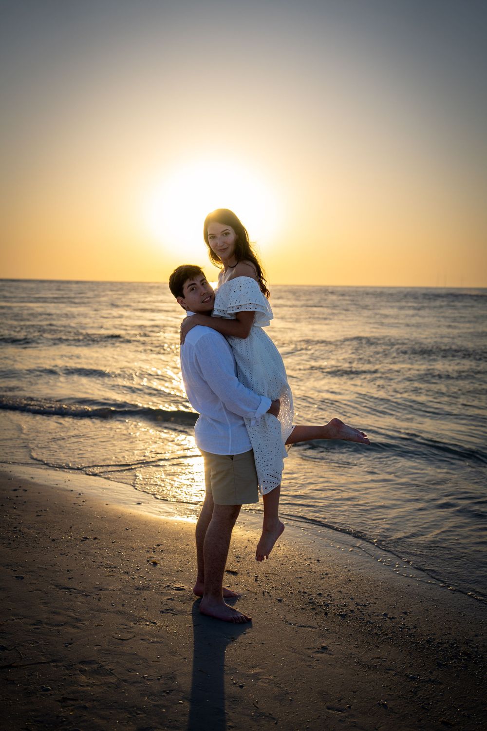 Couple embracing at golden sunset on beach, man lifting woman in white dress near ocean waves in Tampa.