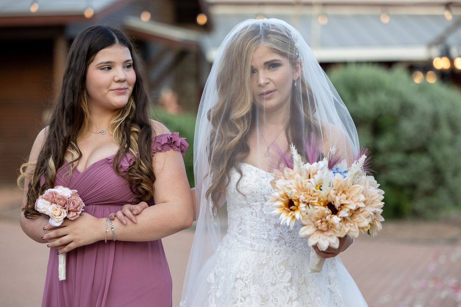 A bride in a white gown and veil stands with her bridesmaid in a mauve dress holding bouquets.